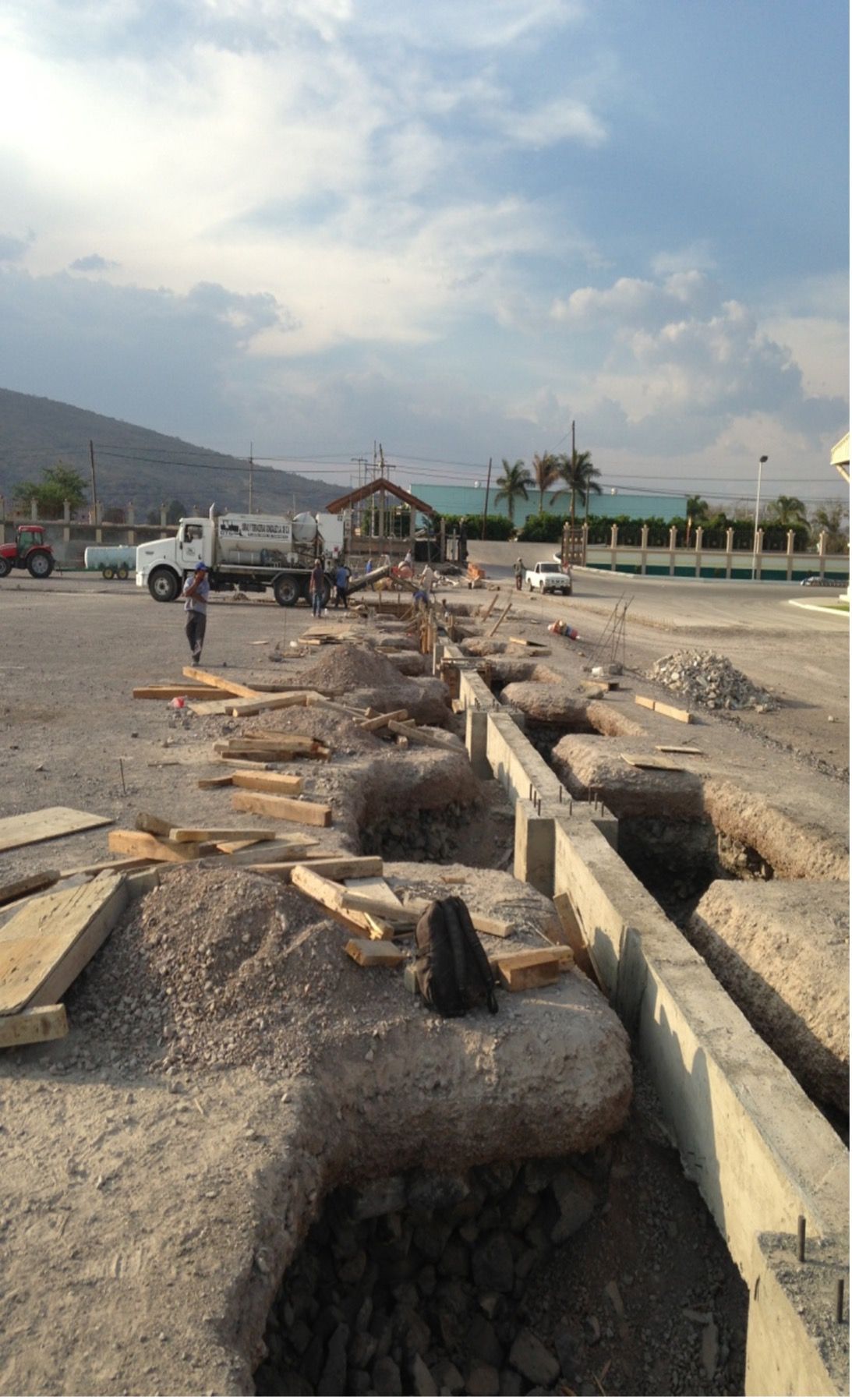Obra en construcción. Zanjas en la grava; un camión y una persona. Montañas y cielo al fondo.