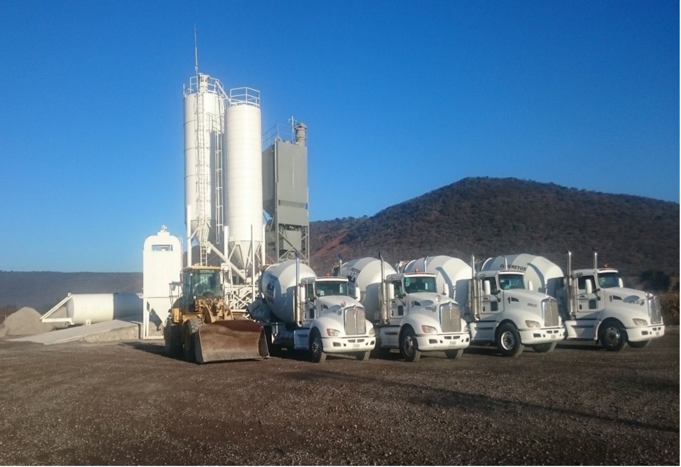 Planta de hormigón con silos, cargadora y camiones hormigonera estacionados en un terreno de grava bajo un cielo despejado.