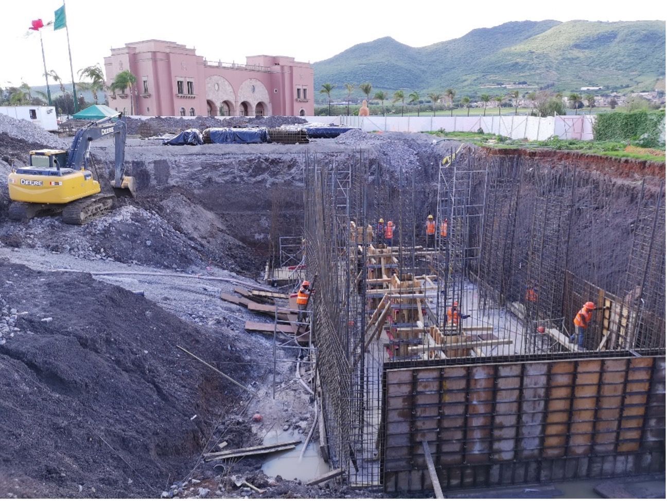 Obra en construcción con obreros, maquinaria y un edificio al fondo.
