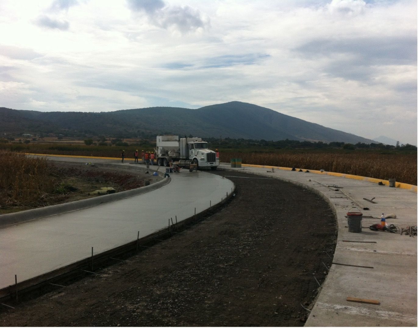 Se vierte hormigón en un tramo curvo de la carretera, con un camión, trabajadores y una montaña al fondo.