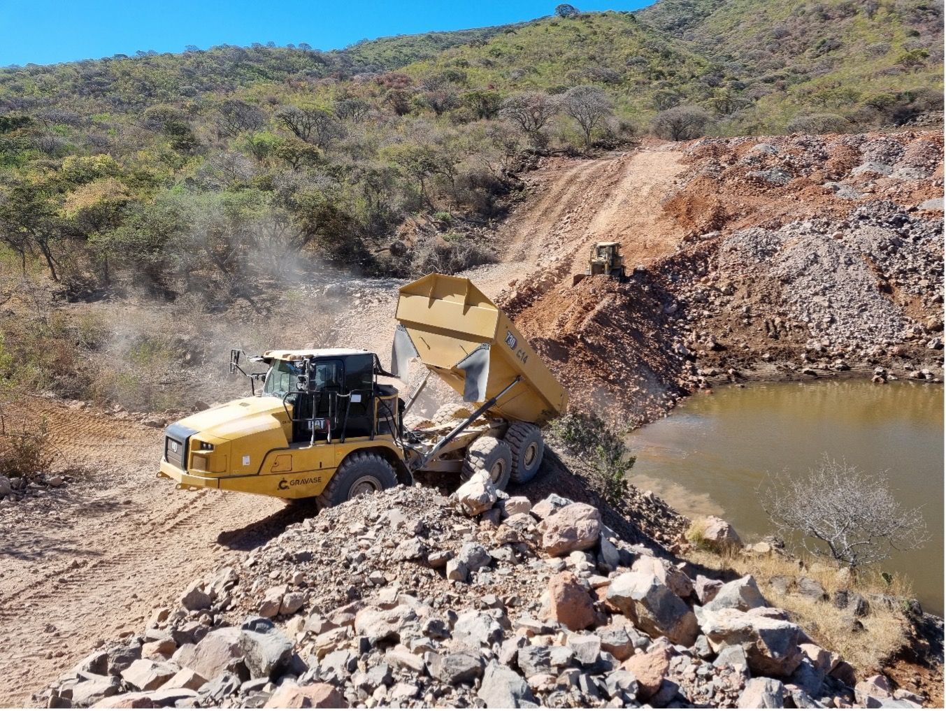 Un camión volquete amarillo descarga rocas cerca de una masa de agua en una ladera.