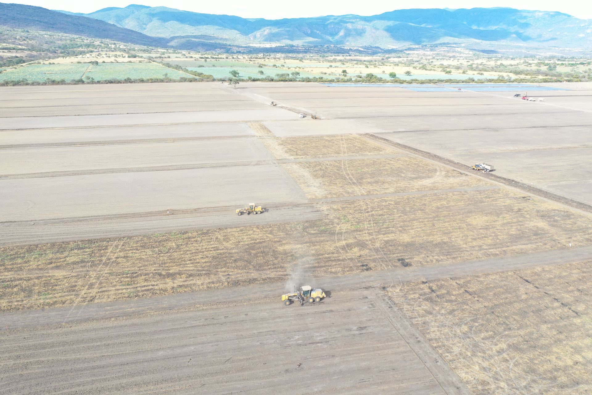 Maquinaria agrícola arando un campo, montañas al fondo bajo un cielo azul.
