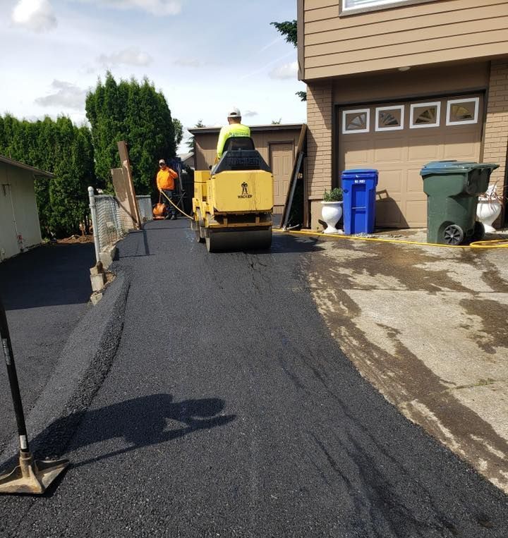 Workers paving a driveway with a roller, near a house and evergreen trees.