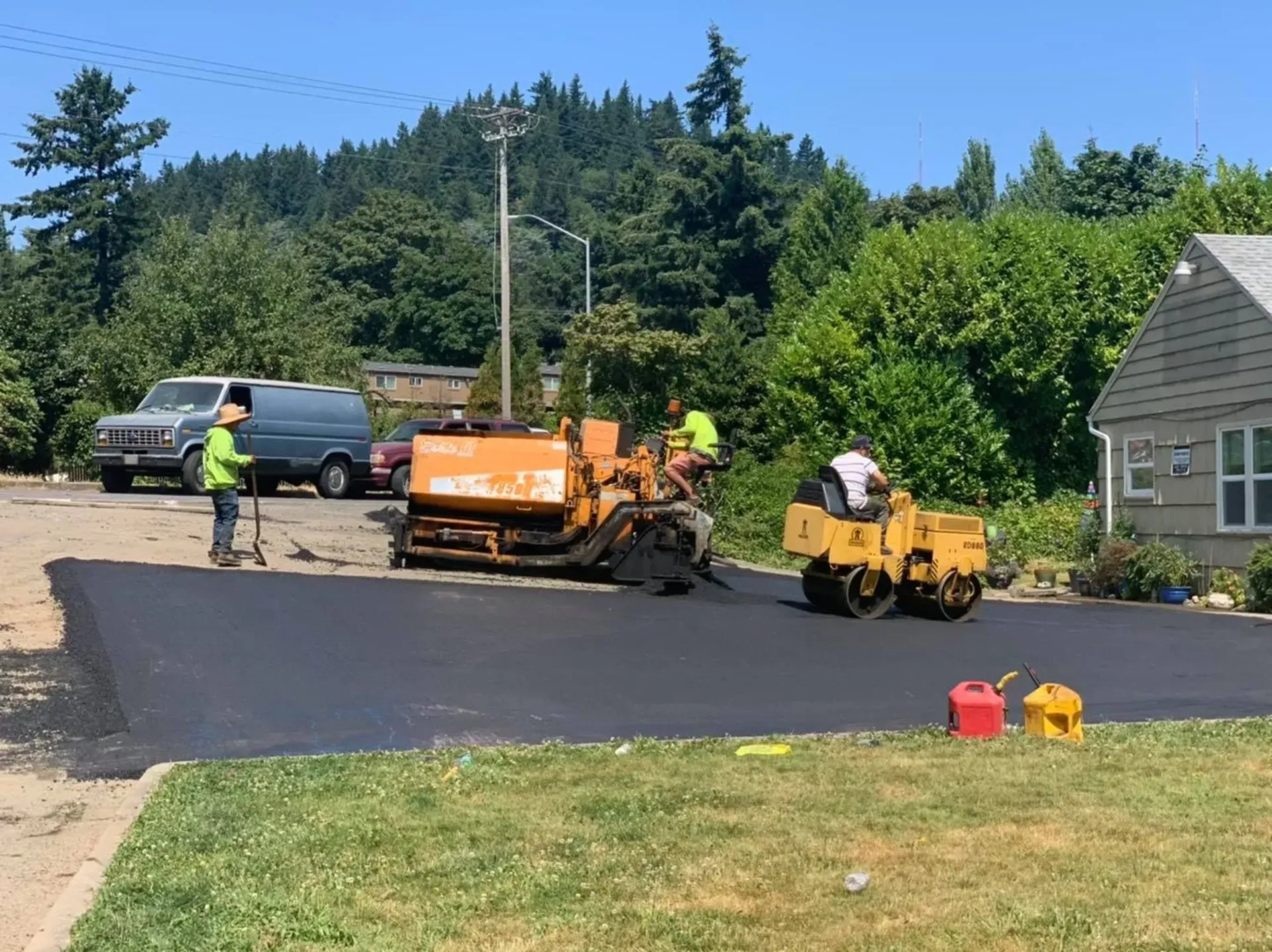 Asphalt paving crew laying fresh blacktop in front of a house, sunny day.