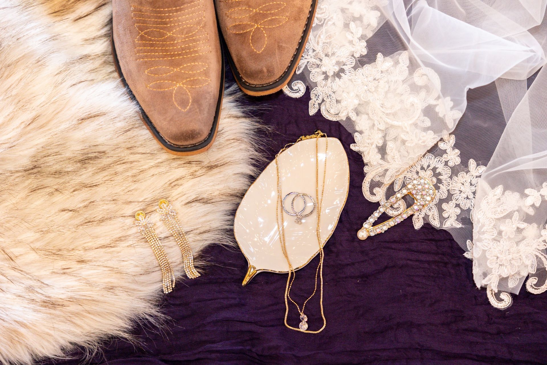 A pair of cowboy boots , a necklace , and a wedding dress are on a purple blanket.