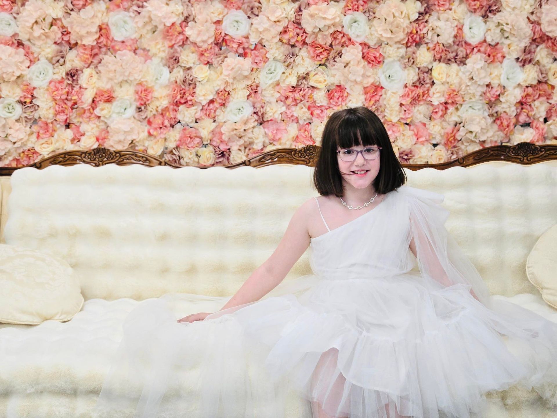 A little girl in a white dress is sitting on a couch.
