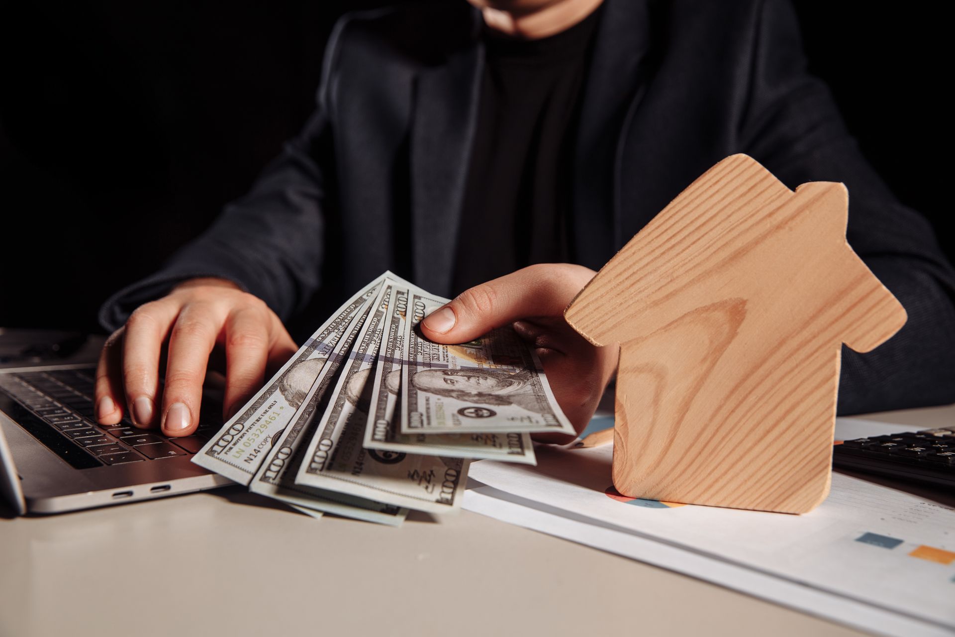 Person holding cash next to a wooden house cutout and laptop on a desk.