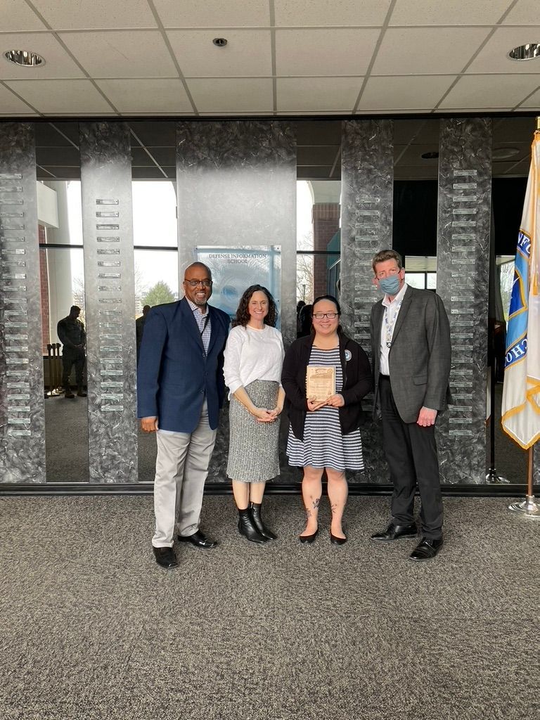 Four people pose in front of a wall with text. Man in a blue jacket, woman in white top, woman with book, and man in a suit.