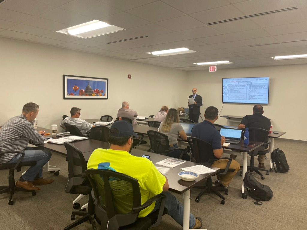A business meeting in a conference room with a presenter standing by a screen and attendees seated at tables.
