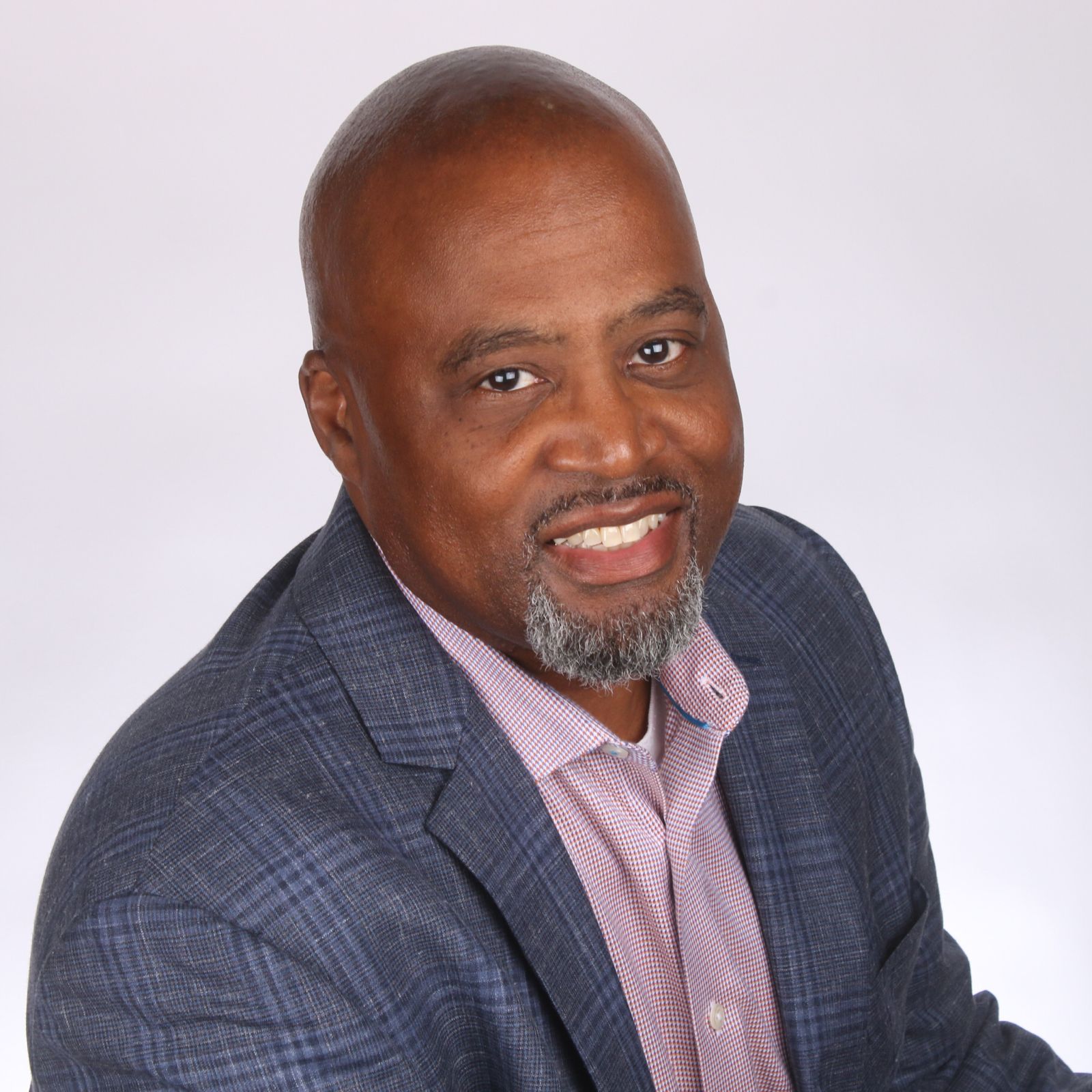 Smiling Black man with a graying beard, wearing a blue plaid blazer and red shirt against a white background.