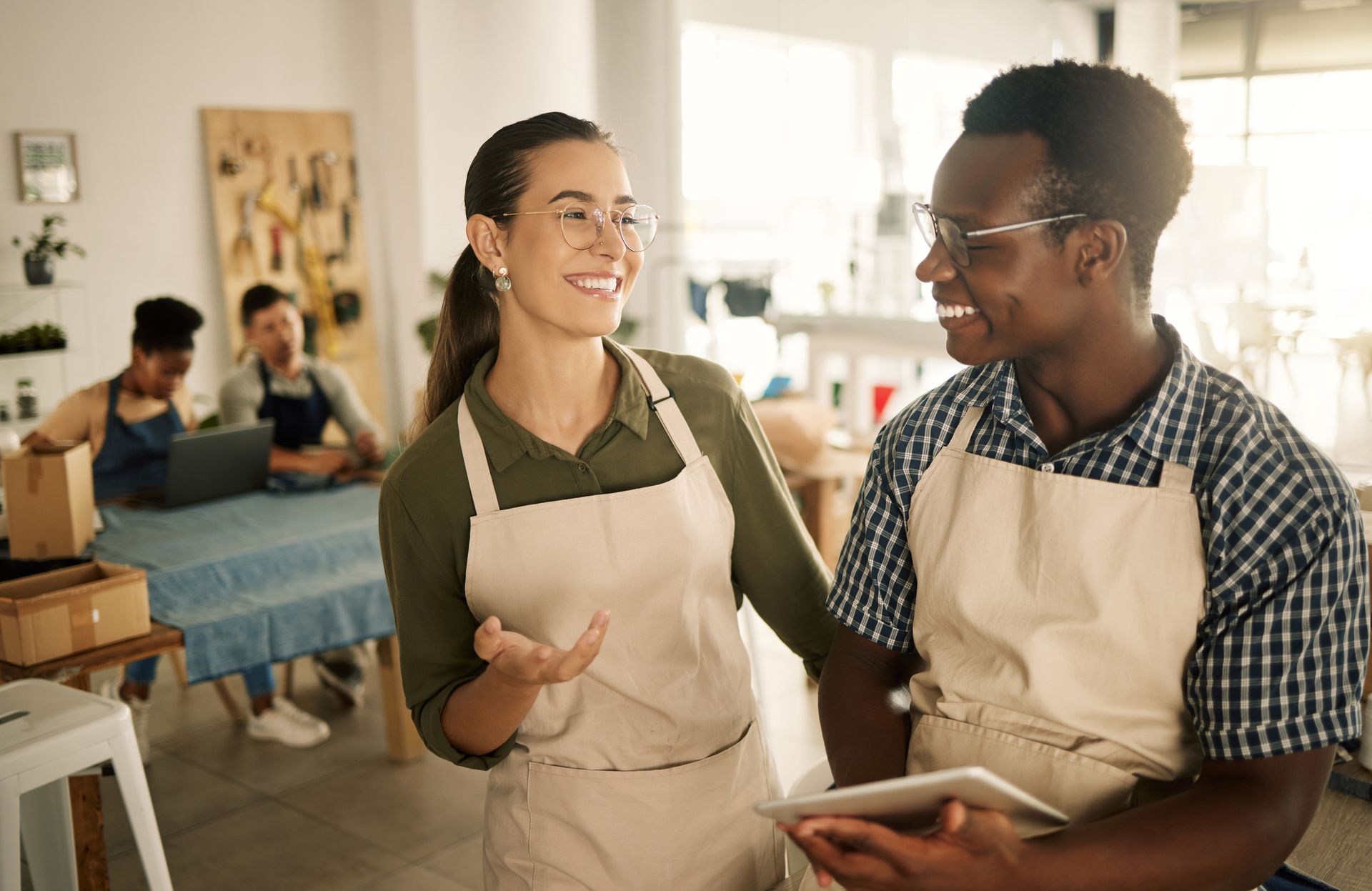 Two people in aprons talking and smiling in a bright workshop, one holding a notebook