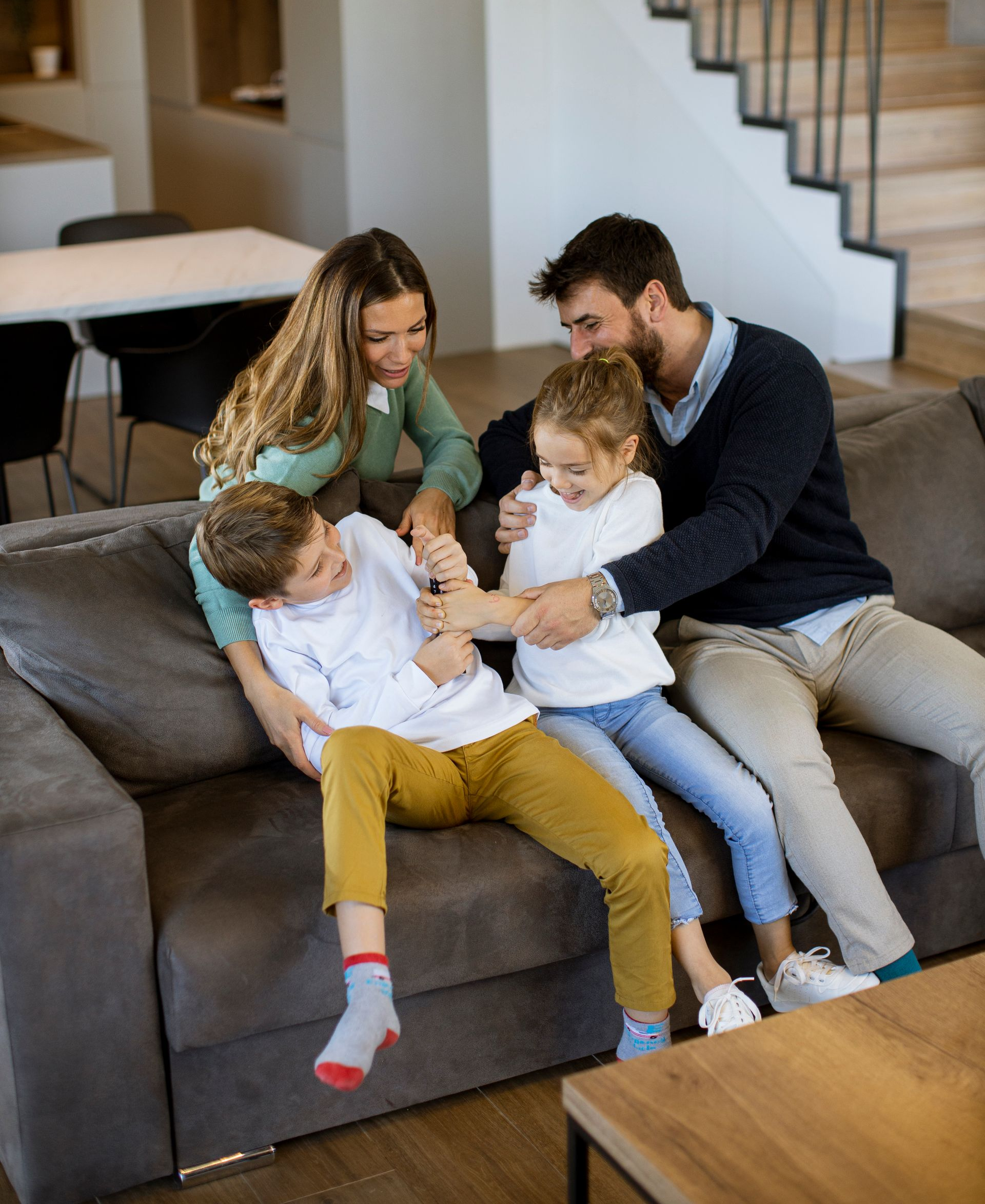 Family of four sitting on a couch, smiling and playing together in a living room.