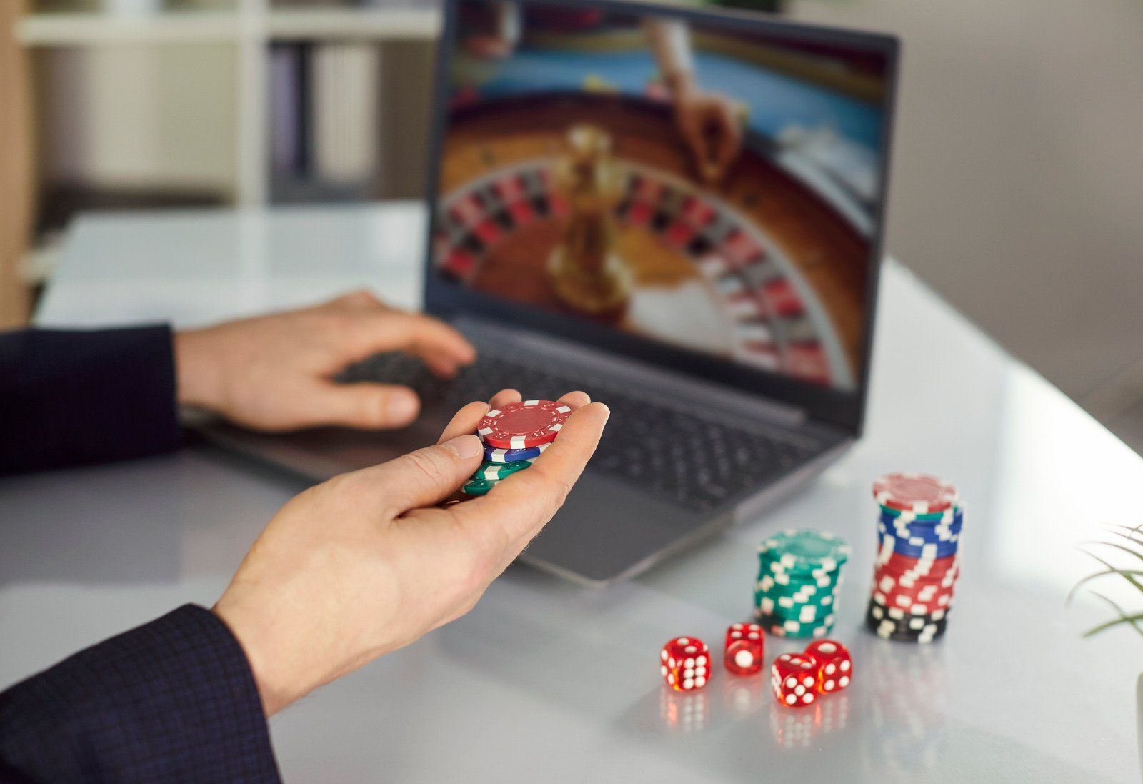 Person playing online roulette on a laptop, holding poker chips, dice and stacks of chips on a desk.