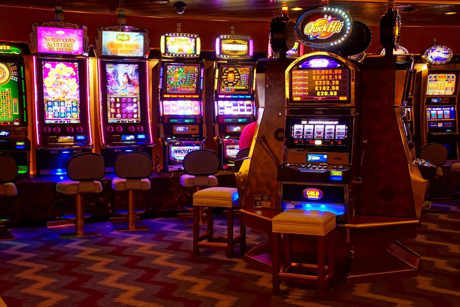 Dimly lit casino with rows of colorful slot machines and empty stools