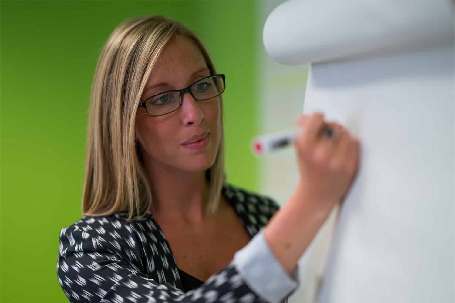 A woman wearing glasses is writing on a whiteboard.