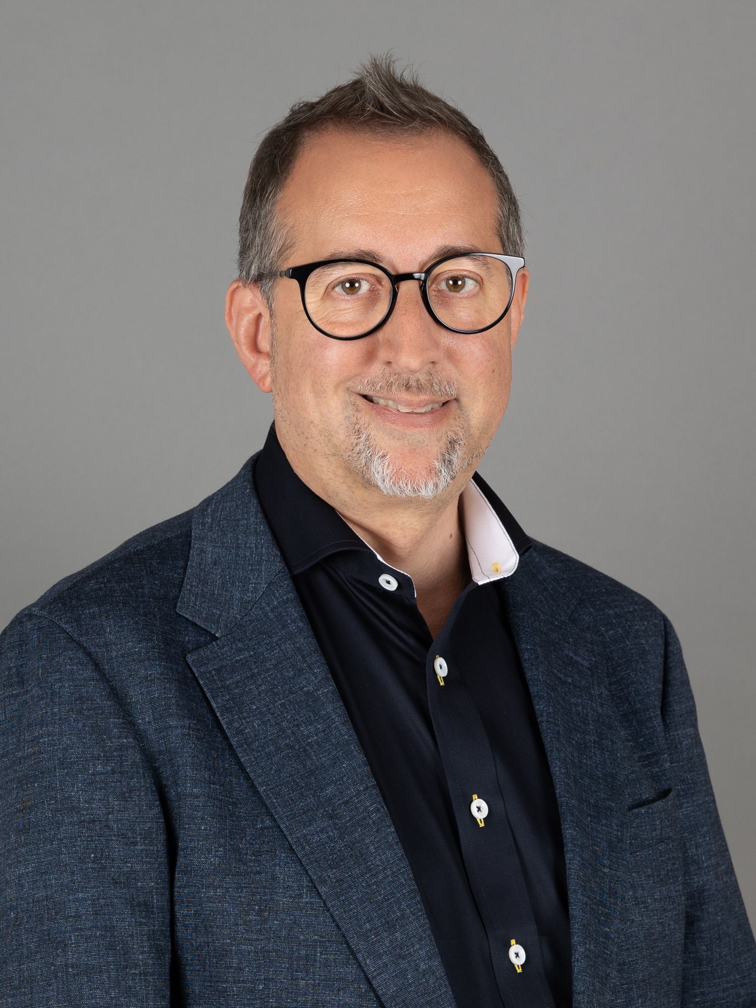 Mann mit Brille, lächelnd, trägt einen blauen Blazer über einem dunkelblauen Hemd vor grauem Hintergrund.