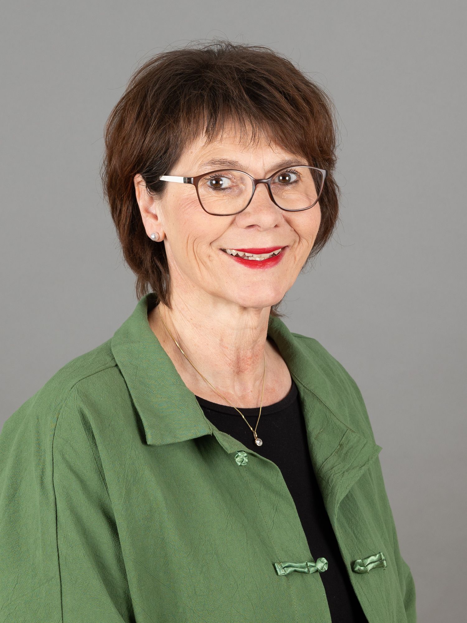 Frau in grüner Jacke und Brille lächelt, grauer Hintergrund.