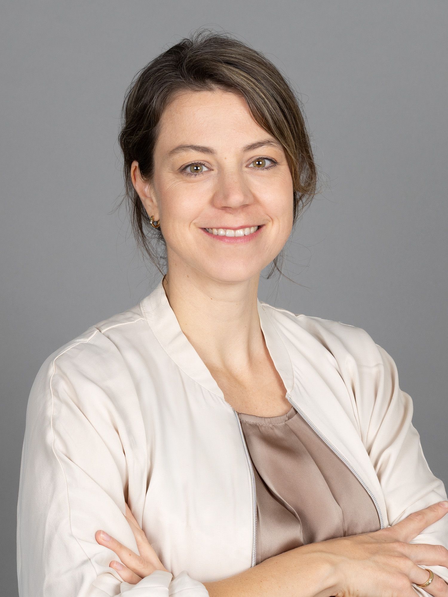 Frau in heller Jacke lächelt mit verschränkten Armen vor grauem Hintergrund.