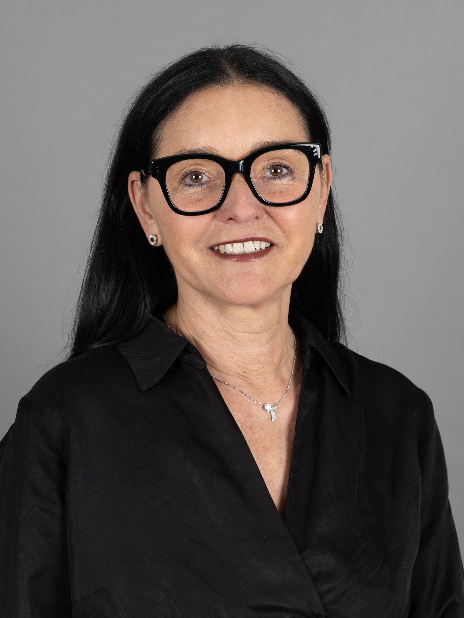 Frau mit Brille und schwarzem Hemd lächelt, den Blick nach vorn gerichtet, vor einem grauen Hintergrund.
