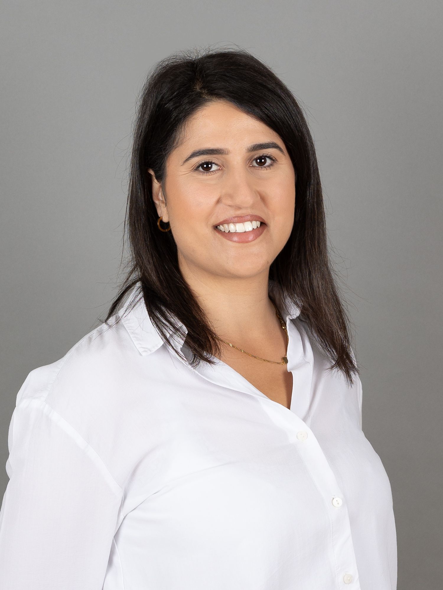 Frau mit dunklen Haaren, die ein weisses Hemd trägt, lächelt vor einem grauen Hintergrund.