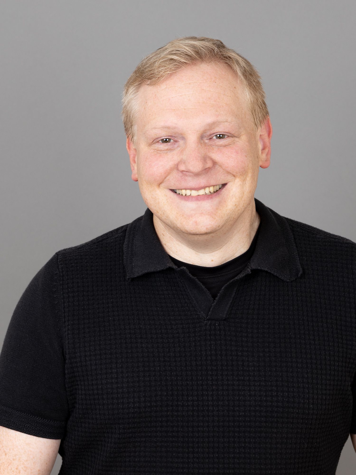 Lächelnde Person mit kurzen, hellen Haaren und schwarzem Hemdkragen vor grauem Hintergrund.