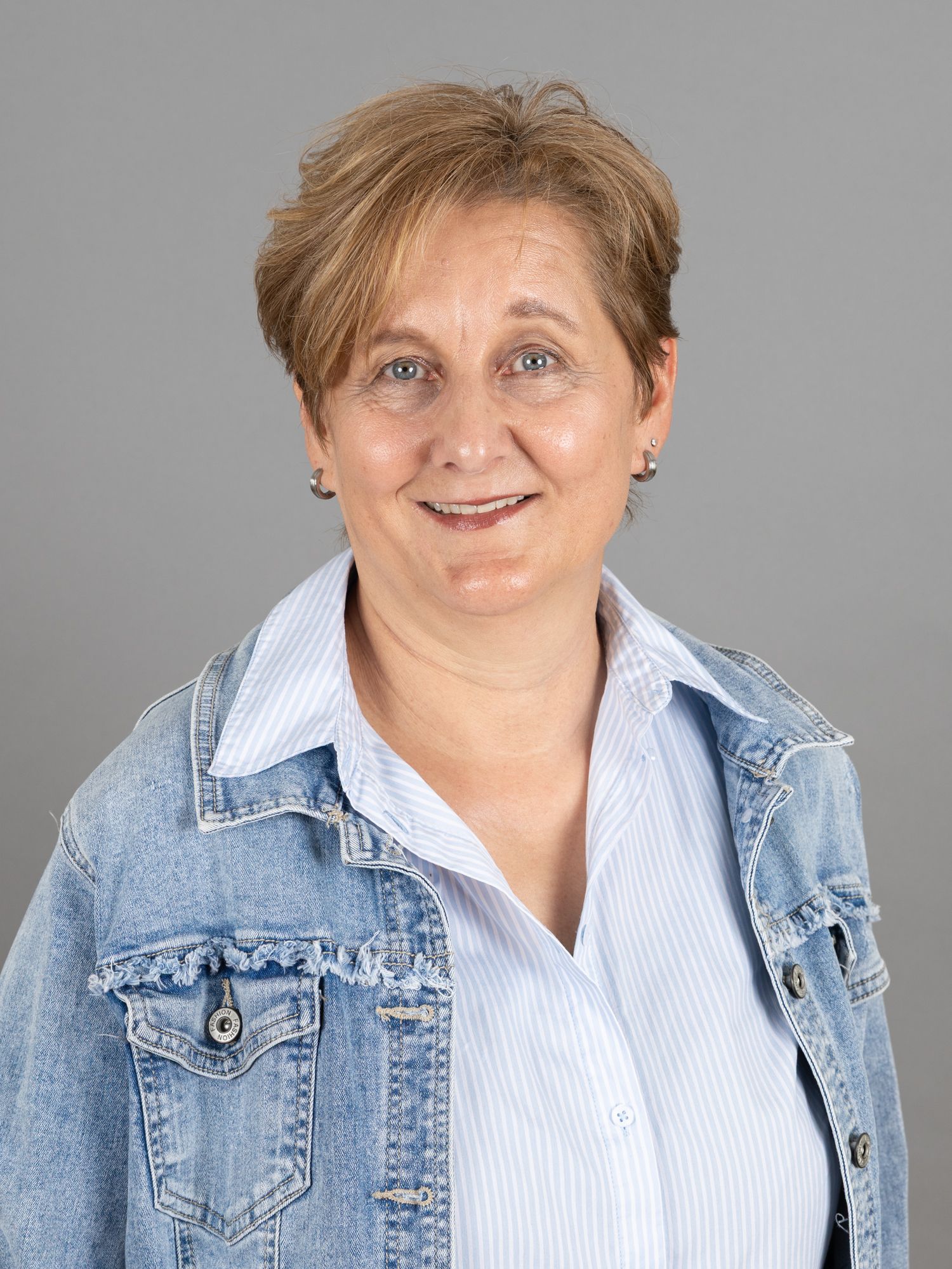 Frau mit kurzen braunen Haaren, die eine Jeansjacke über einem hellblau gestreiften Hemd trägt, lächelt.