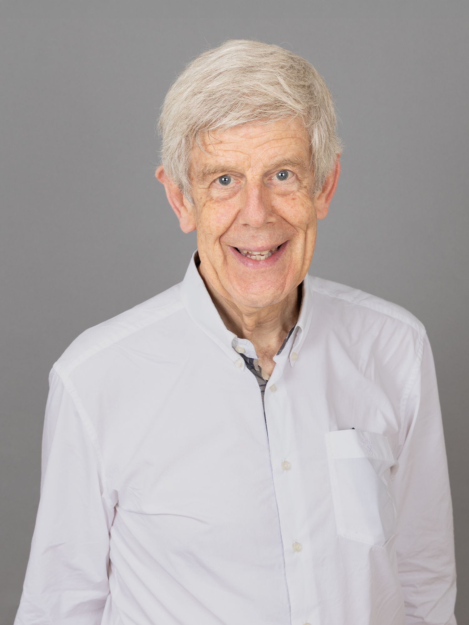 Lächelnder Mann mit weissen Haaren, der ein weisses Hemd mit Knöpfen trägt, vor grauem Hintergrund.