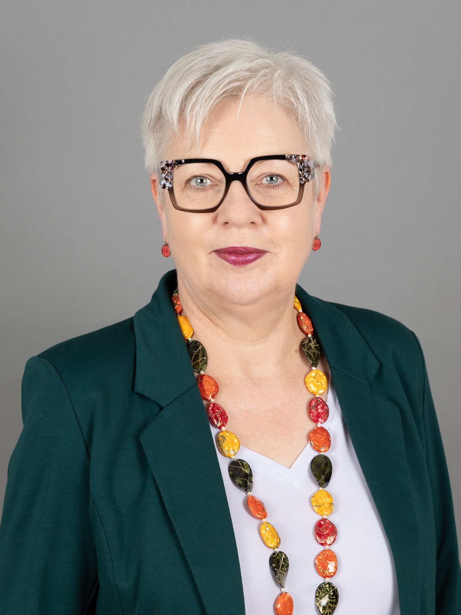 Frau mit kurzen weissen Haaren, Brille und bunter Halskette, die einen grünen Blazer und ein weißes Hemd trägt.