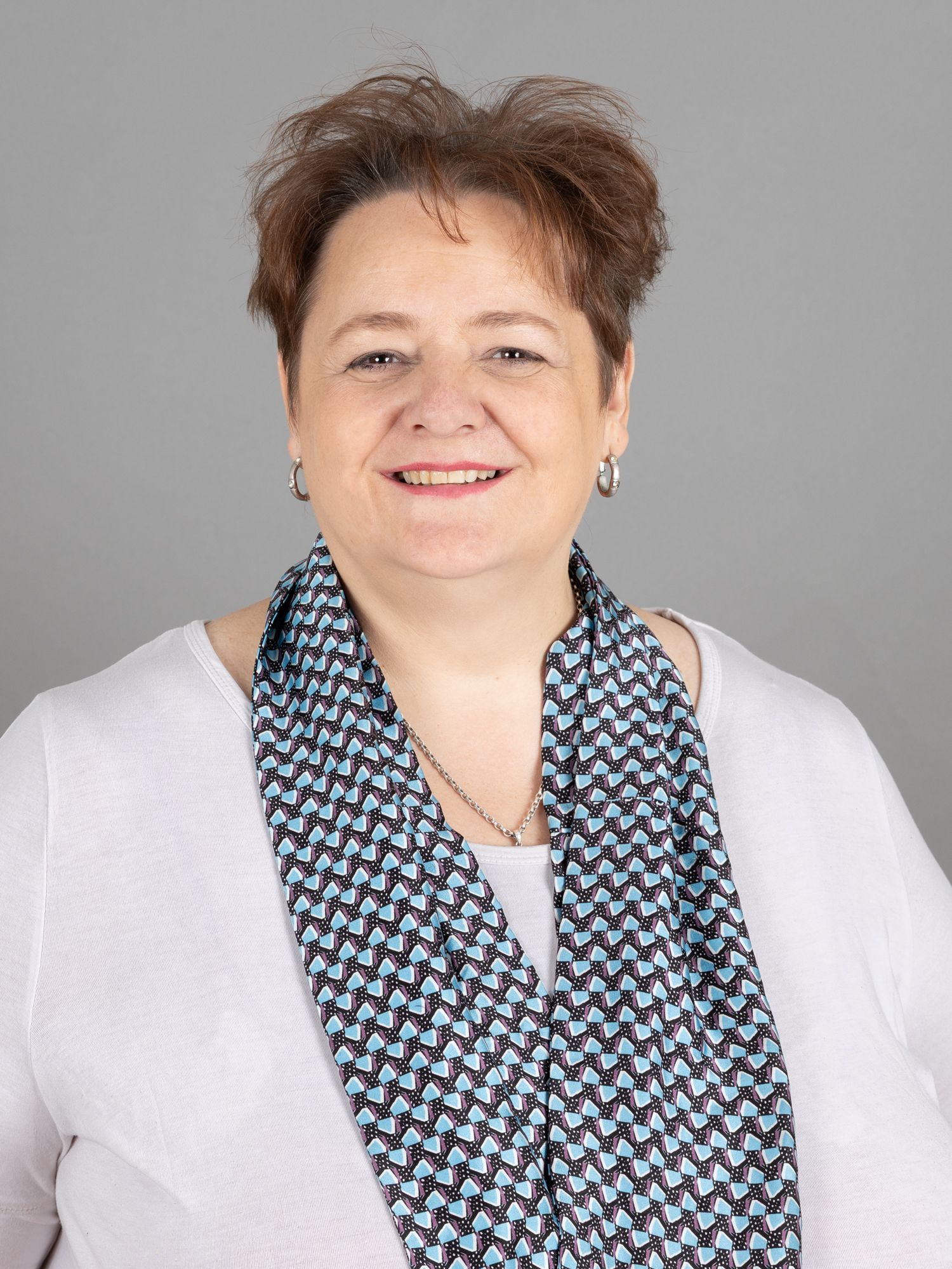 Eine Frau mit kurzen braunen Haaren lächelt. Sie trägt einen gemusterten blau-weissen Schal und ein weißes Hemd.