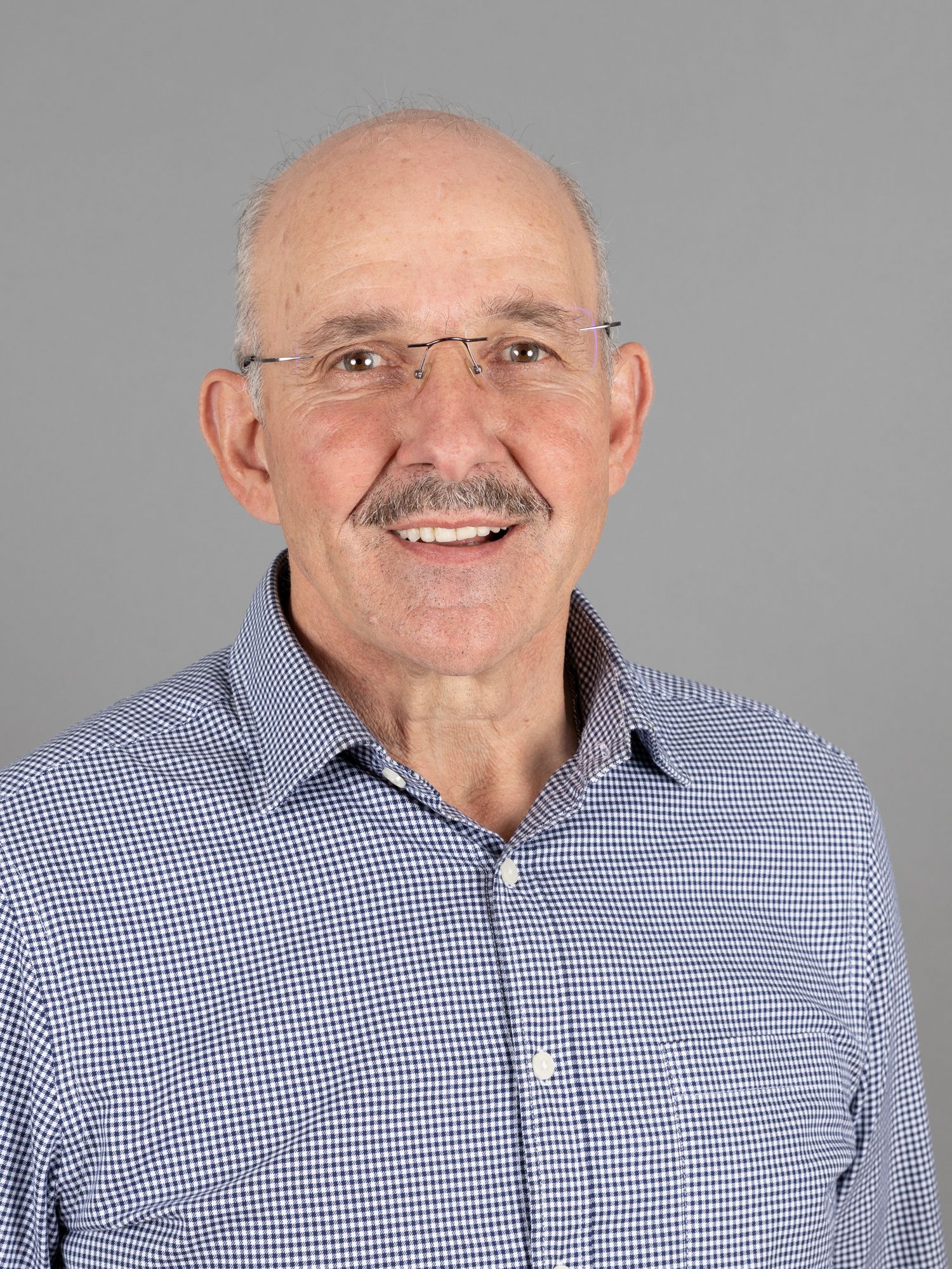 Ein Mann mit Brille und Schnurrbart lächelt vor einem grauen Hintergrund. Er trägt ein blau gemustertes Button-Down-Hemd.