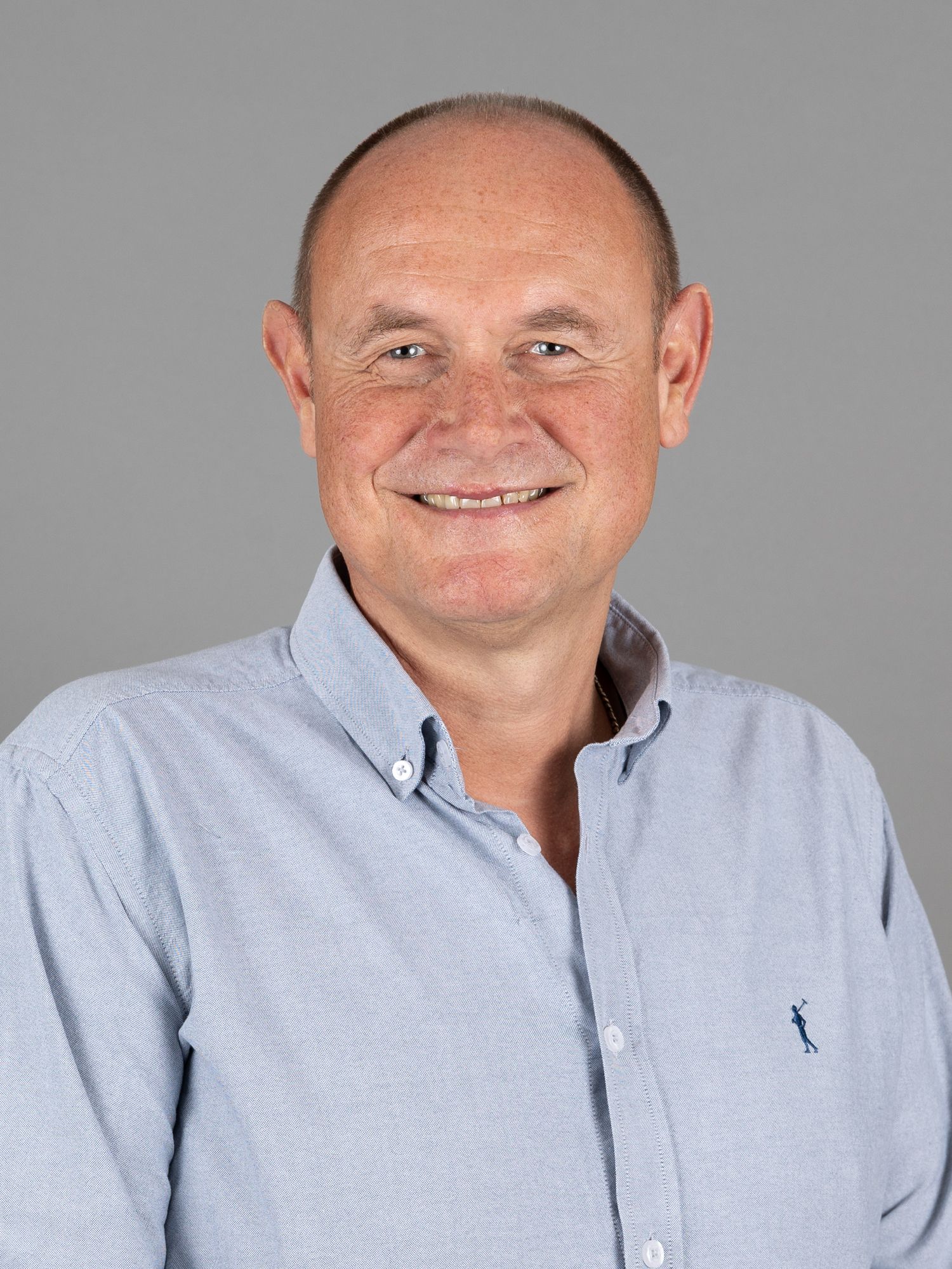 Mann mit hellblauem Button-Down-Hemd lächelt vor grauem Hintergrund.