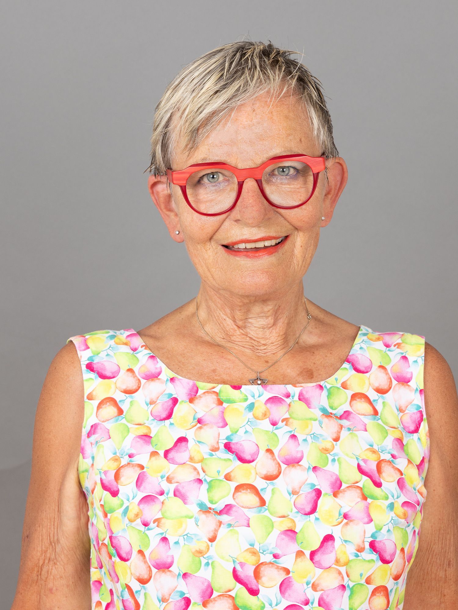 Frau mit roter Brille und geblümtem Kleid lächelt vor grauem Hintergrund.