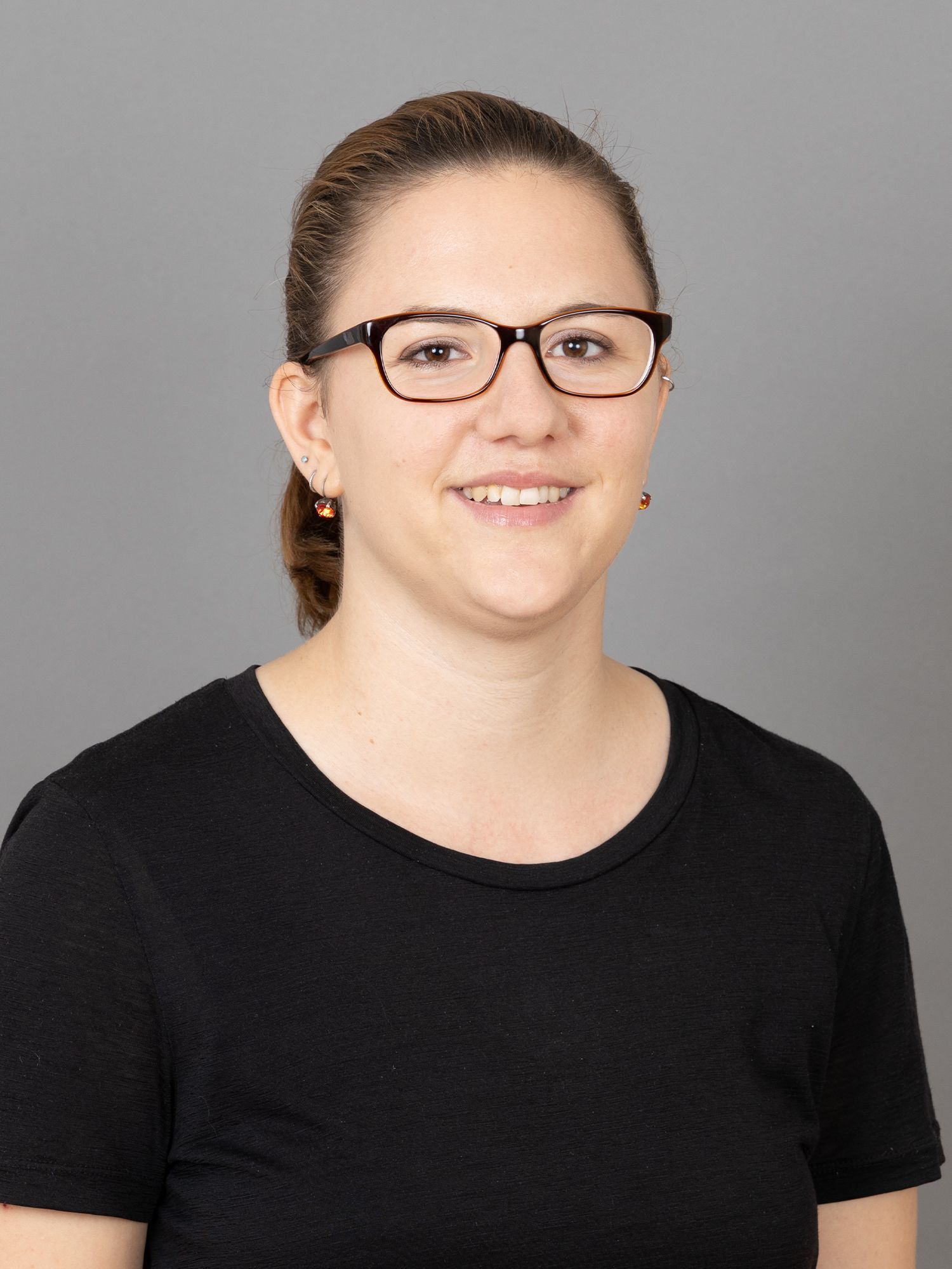 Frau mit Brille lächelt, trägt ein schwarzes Hemd vor grauem Hintergrund.