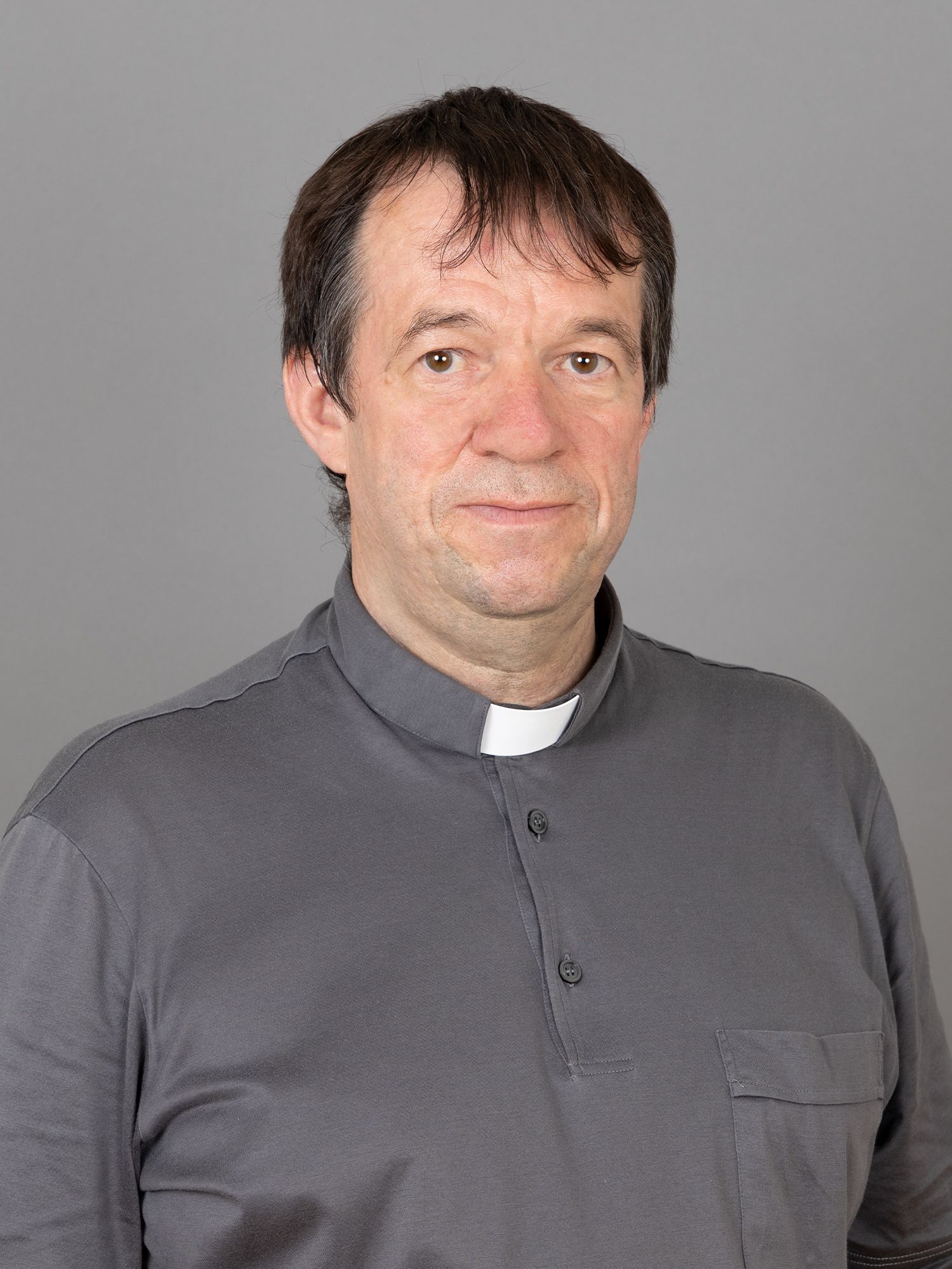 Mann im Priesterkragen, graues Hemd, lächelt vor grauem Hintergrund.
