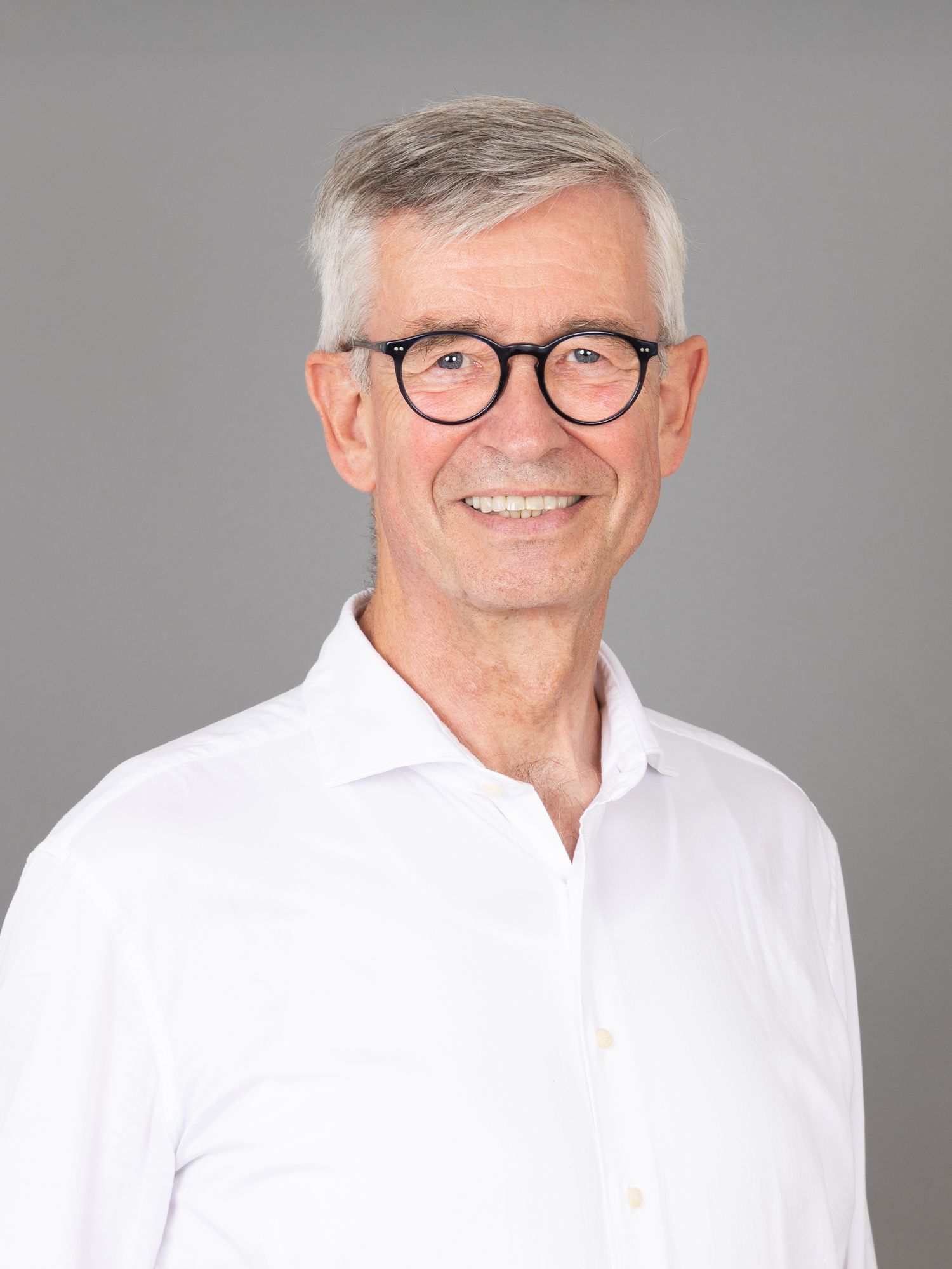 Mann mit Brille lächelt, trägt ein weisses Hemd vor grauem Hintergrund.
