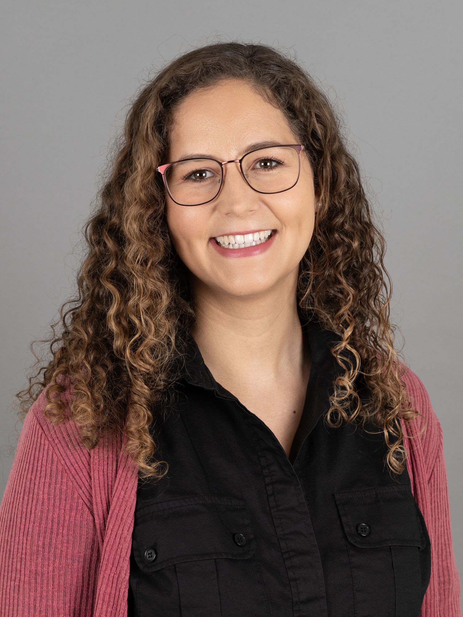 Frau mit lockigem braunem Haar, Brille, lächelnd, trägt ein schwarzes Hemd und eine rosa Strickjacke vor grauem Hintergrund.
