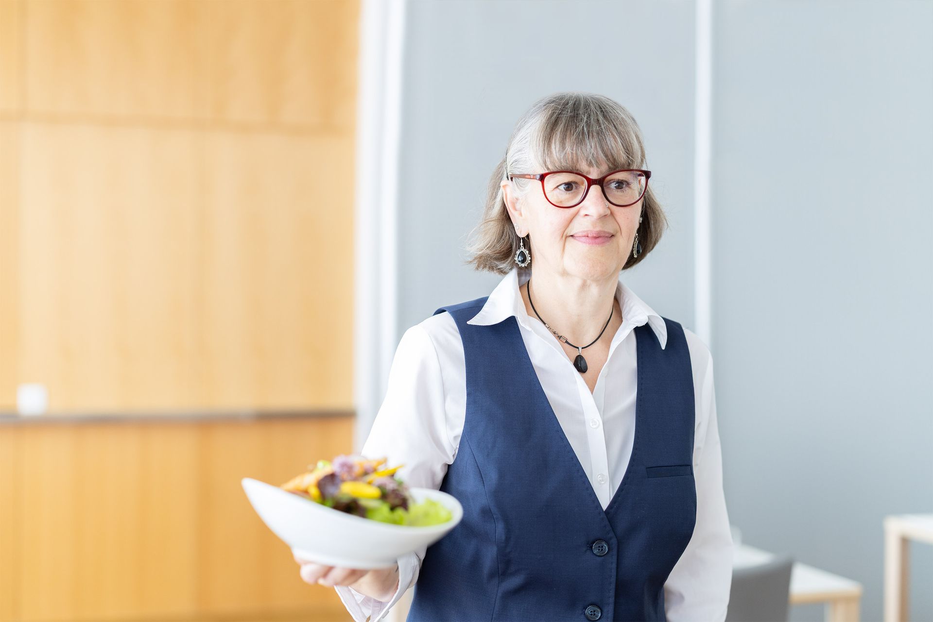 Frau in blauer Weste, die eine Schüssel Salat hält, lächelt. Weisses Hemd, Brille und graues Haar.