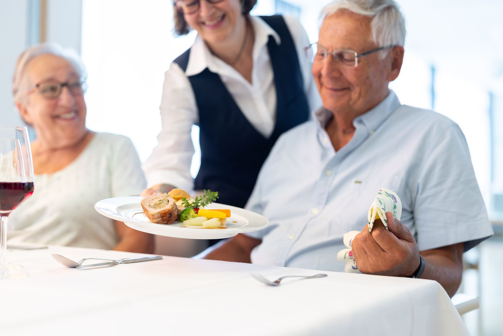 Eine Kellnerin serviert einem älteren Paar am Esstisch ein auf einem Teller angerichtetes Essen.
