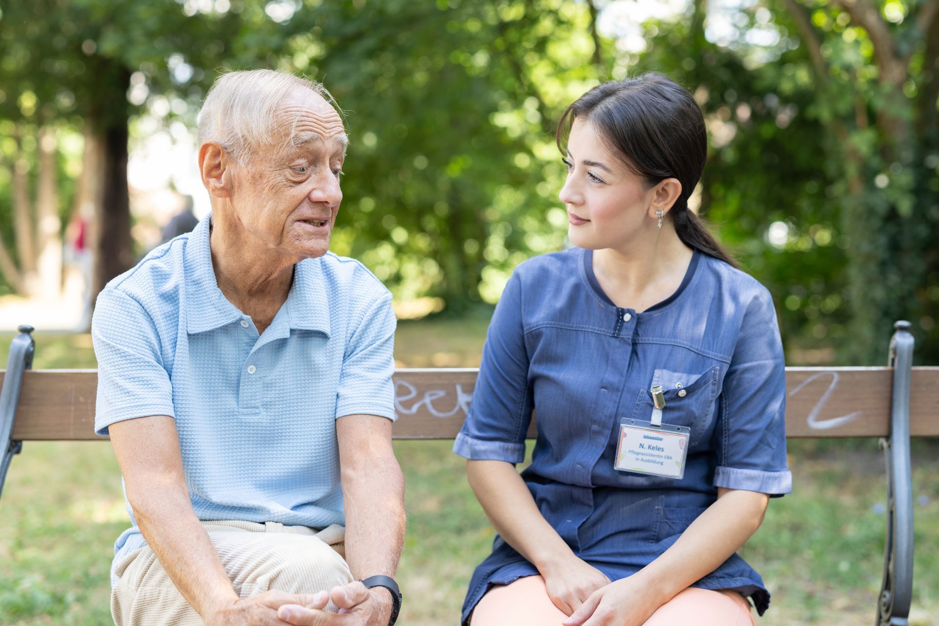 Ältere Person und Pflegekraft sitzen auf einer Parkbank und unterhalten sich. Grüner Hintergrund.