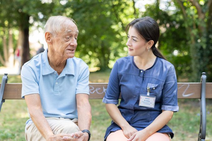 Ältere Person und Pflegekraft sitzen auf einer Parkbank und unterhalten sich. Grüner Hintergrund.