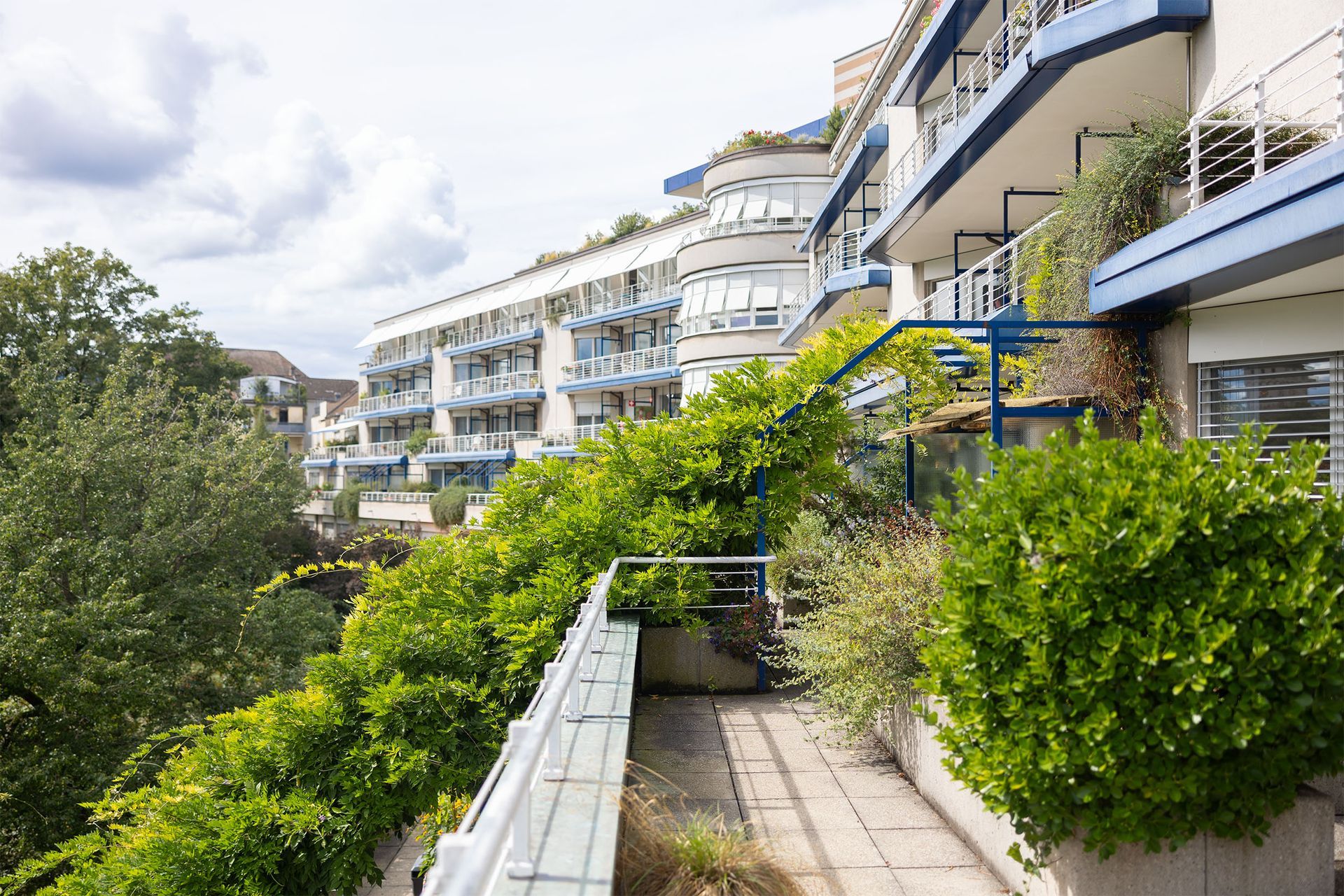 Aussenansicht eines mehrstöckigen Gebäudes mit Balkonen, Grünflächen und blauen Akzenten. Bewölkter Himmel.