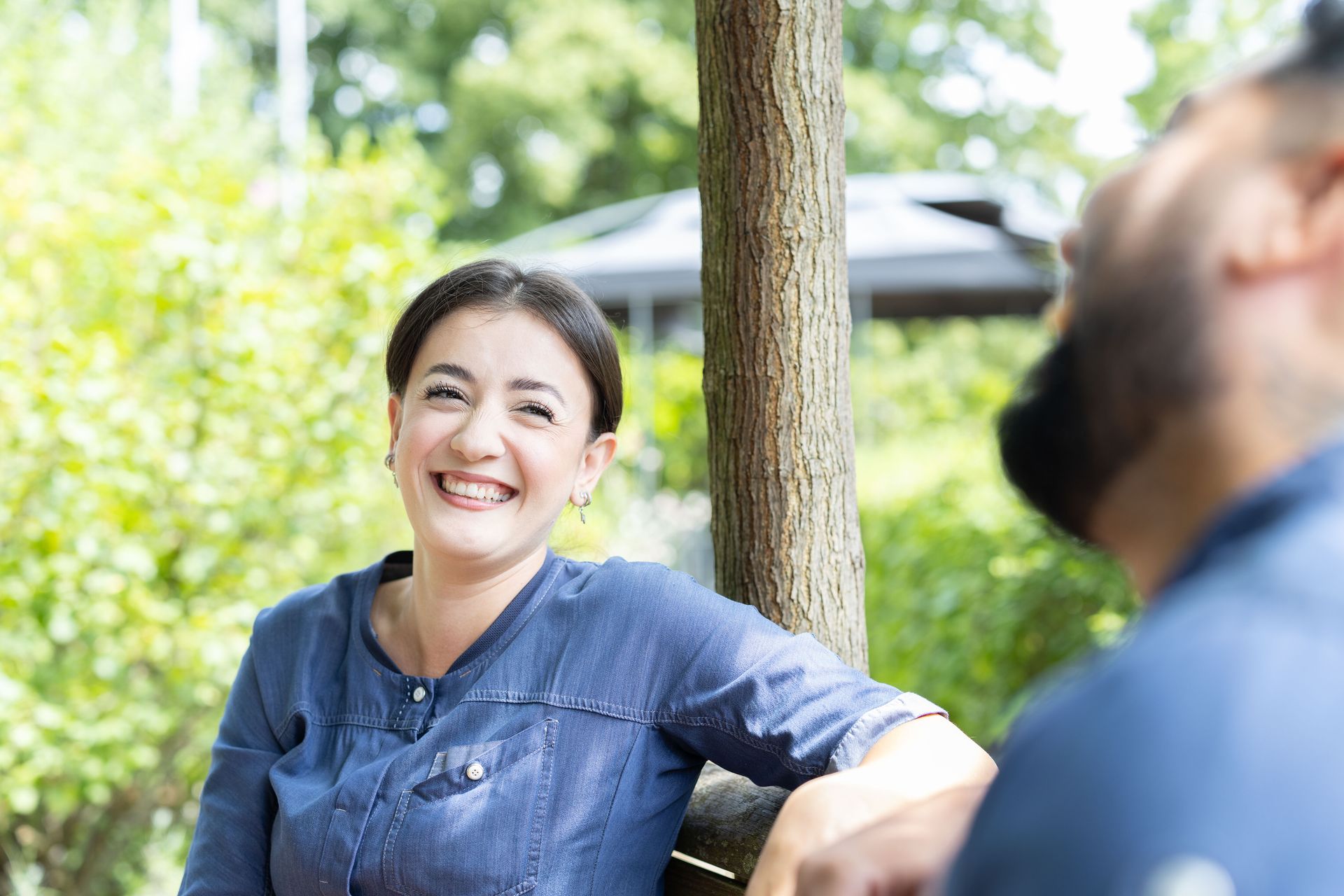 Eine lächelnde Frau lehnt sich an eine Bank und blickt zu einer unscharfen Person in einer Gartenlandschaft.