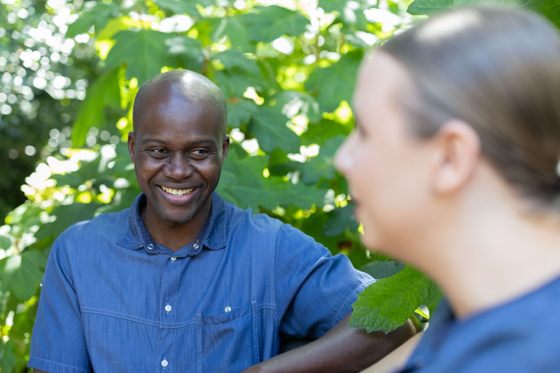 Ein Mann in blauem Hemd lächelt und unterhält sich mit einer Person, die teilweise im Bild zu sehen ist; im Hintergrund ist Grün zu sehen.