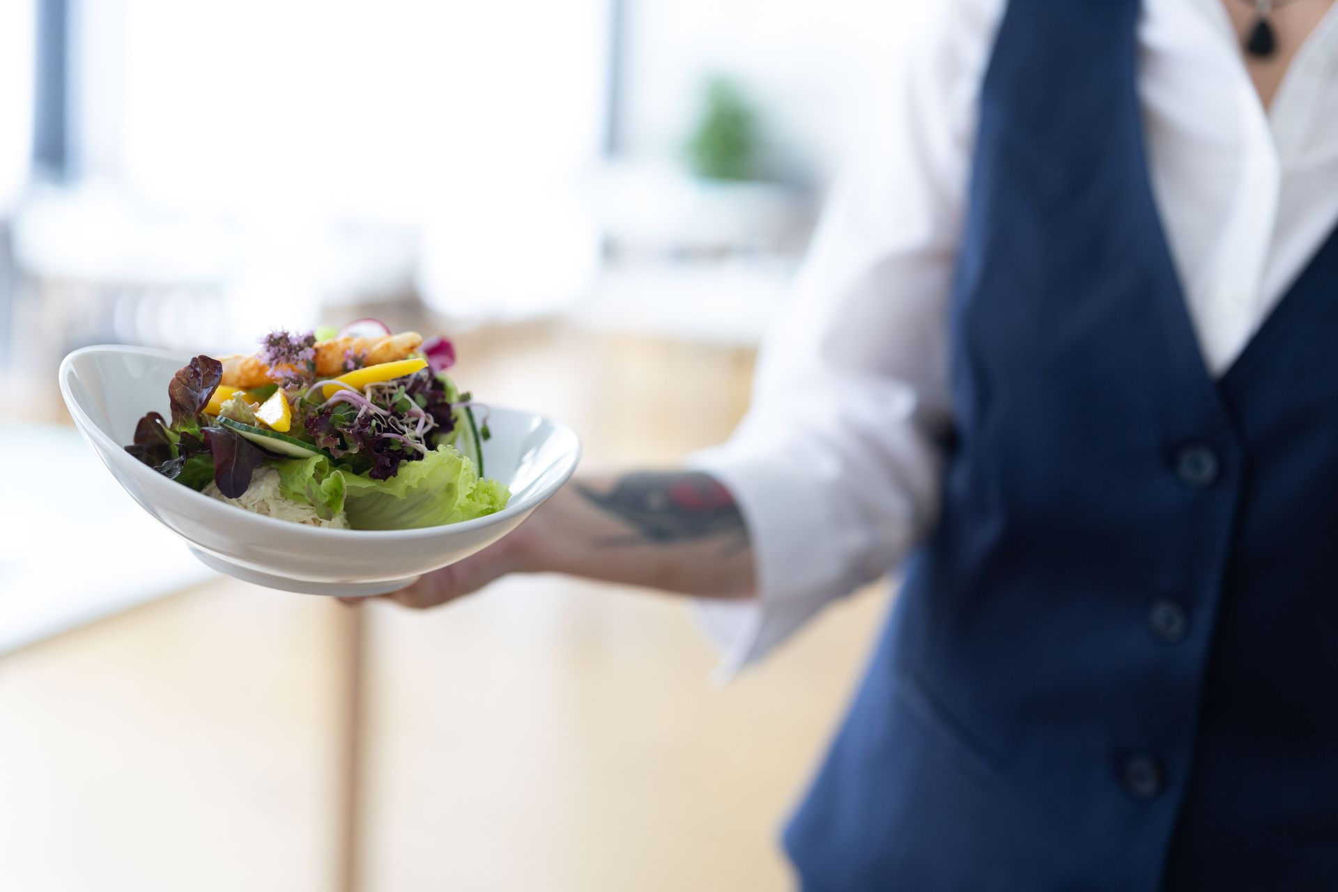 Kellner mit einer weissen Schüssel Salat; trägt eine blaue Weste und ein weisses Hemd; drinnen.