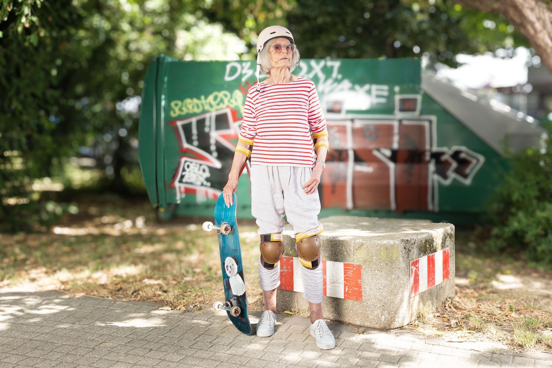 Eine Person in Schutzausrüstung hält ein Skateboard und steht neben einem mit Graffiti besprühten grünen Mülleimer und einer Betonbarriere.