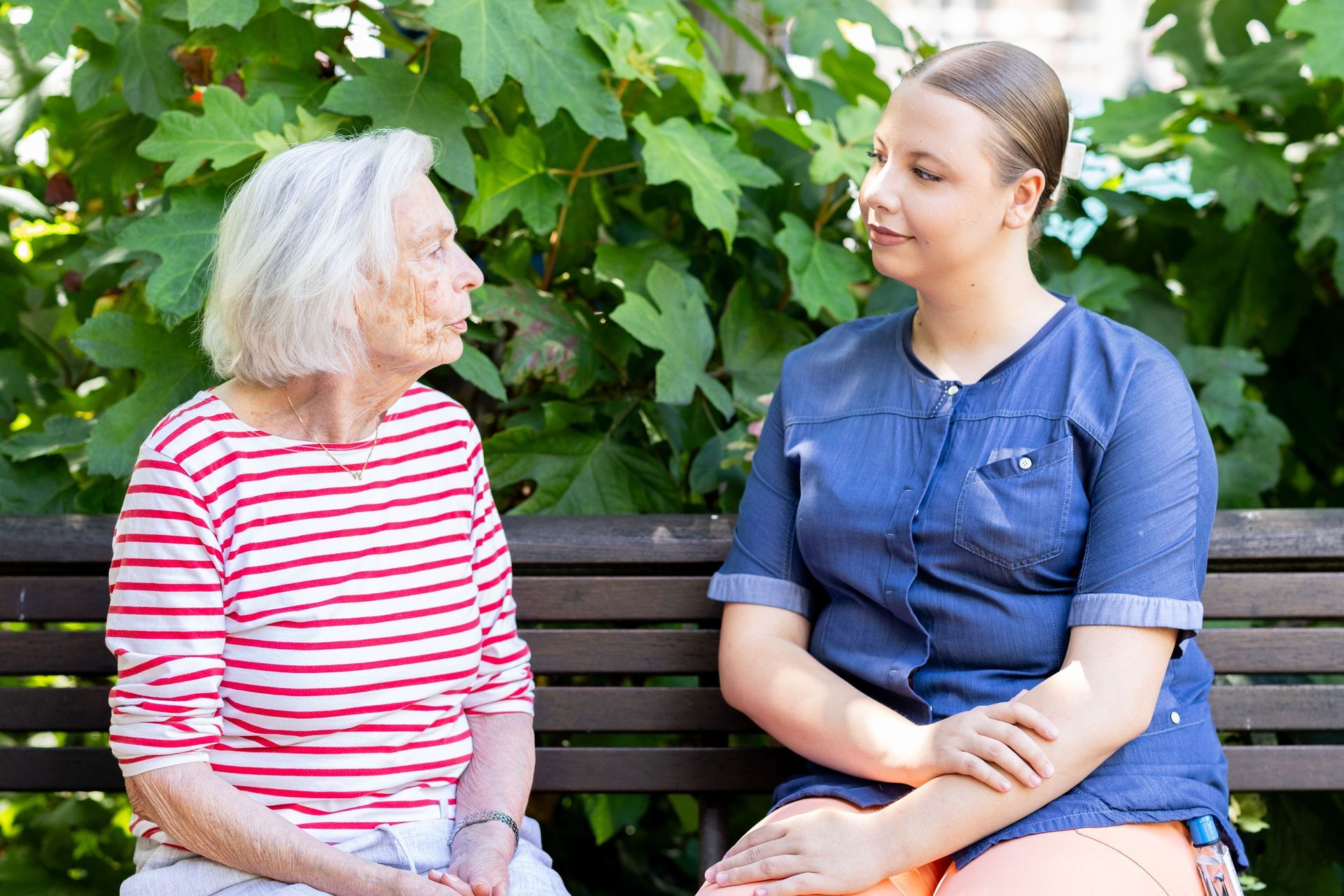 Eine ältere und eine jüngere Frau sitzen auf einer Parkbank und unterhalten sich. Im Hintergrund ist grünes Laubwerk zu sehen.