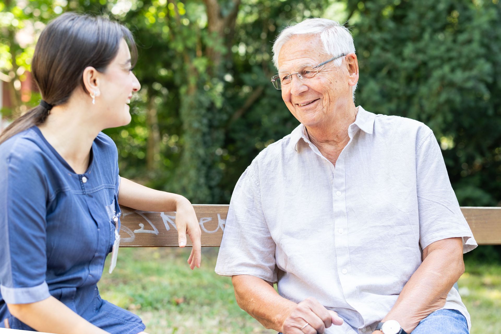Eine Frau in Blau und ein älterer Mann in weissem Hemd sitzen lächelnd auf einer Bank in einem Park.