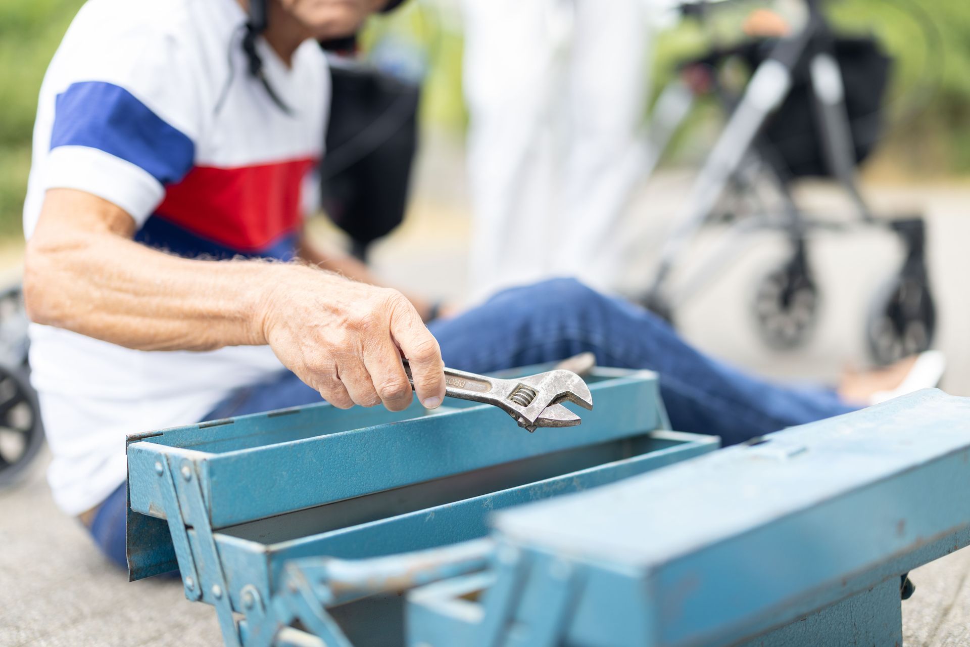 Ein Mann sitzt mit einem Werkzeugkasten auf dem Boden, neben ihm ein Rollator. Er hält einen Schraubenschlüssel.