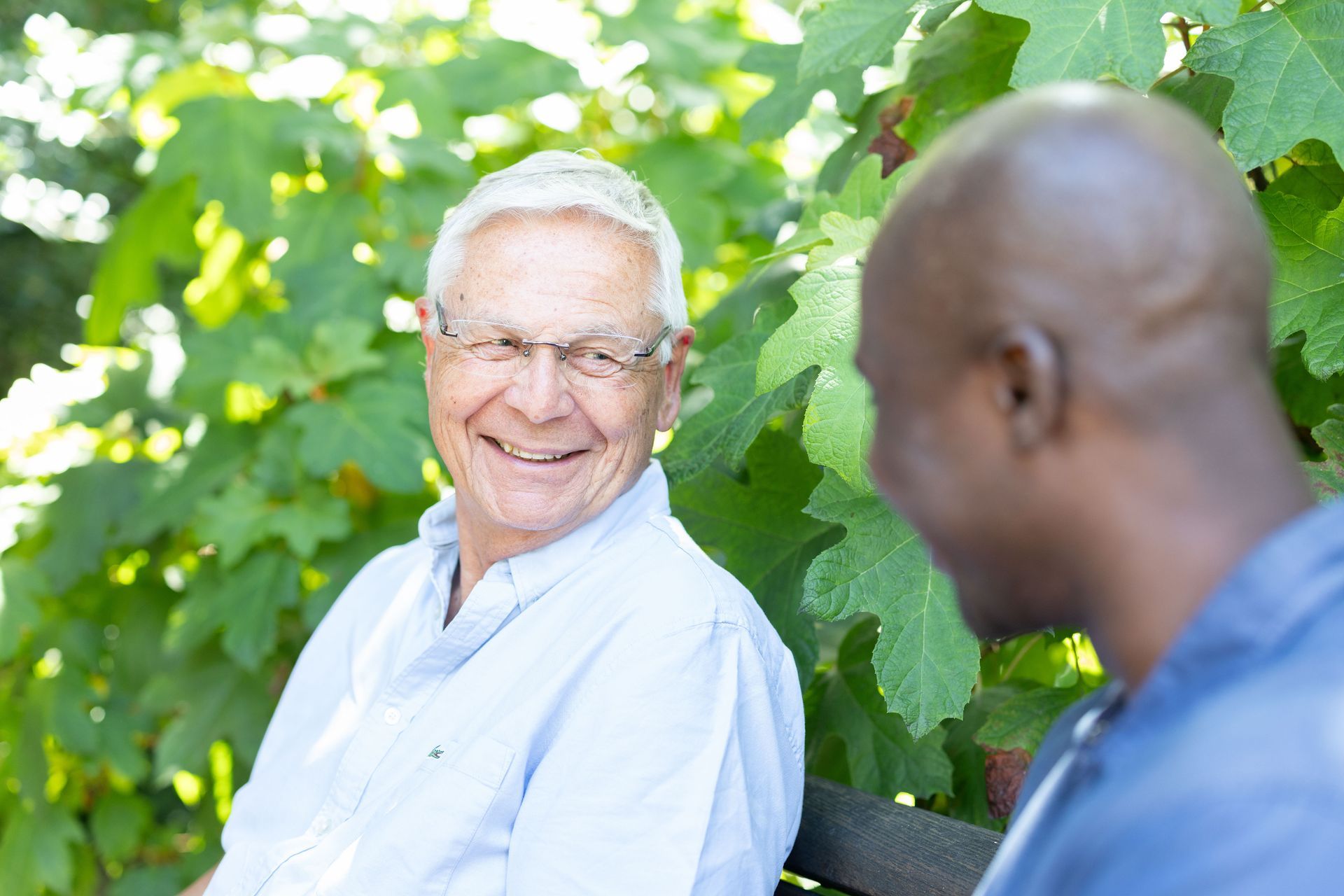 Ein älterer Mensch mit Brille lächelt im Gespräch mit einer Person, die auf einer Bank inmitten von Grün sitzt.