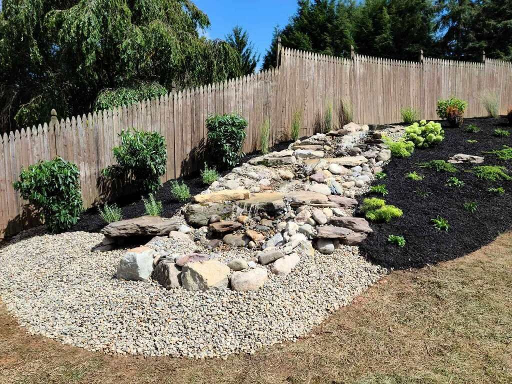 There is a waterfall in the backyard with a wooden fence in the background.